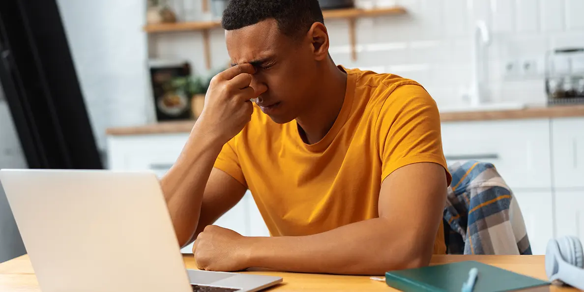 Young man in a yellow shirt sitting at a desk in front of a laptop, pressing his fingers to the bridge of his nose in apparent pain