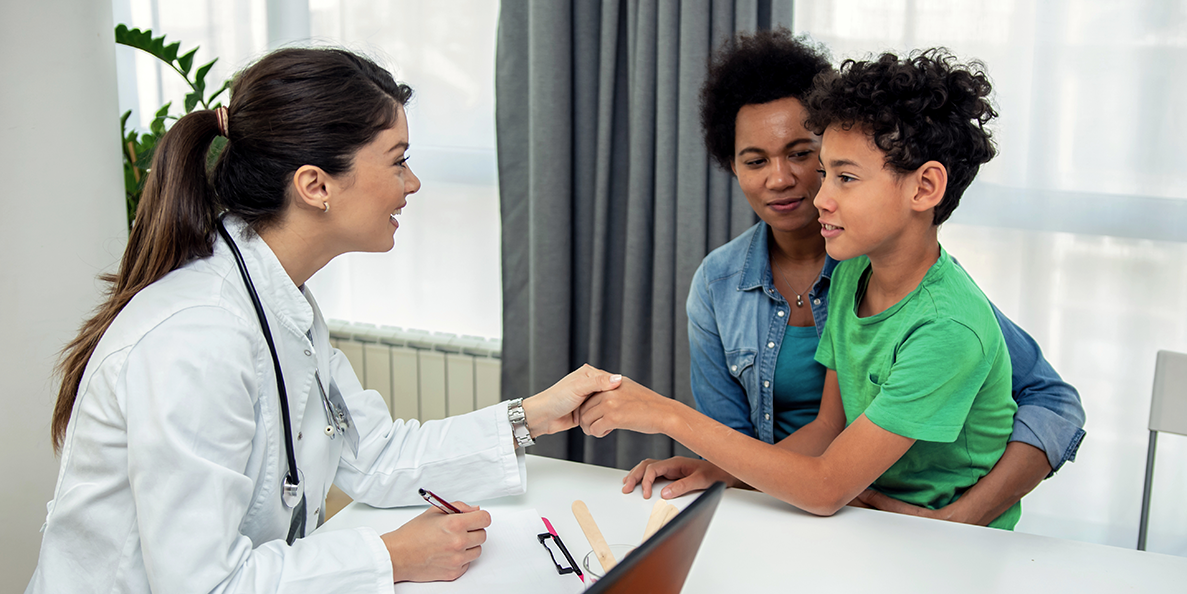Healthcare professional in a white lab coat shaking hands with a young patient across a desk, while an adult seated behind the child watches supportively